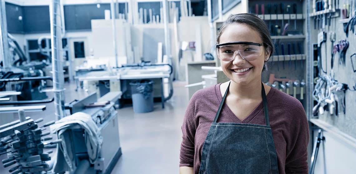 Auszubildende mit Arbeitsschürze und Schutzbrille lächelnd in Werkstatt #Partnerschaftliche Ausbildung 